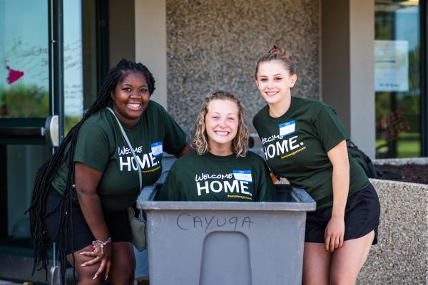 A Laker Move-In Crew member sitting in a gray move-in bin, with two members standing on each side of the bin, all smiling at the camera