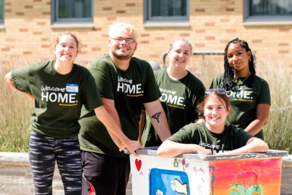 Four Laker Move-In Crew members standing side by side behind a move-in bin that another member is sitting in, all smiling at the camera