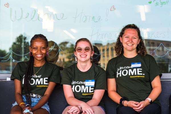 Three Laker Move-In Crew members smiling at the camera, sitting in front of a Waterbury Hall window. The message "Welcome Home! Home of the Lakers" is written on the window with window markers