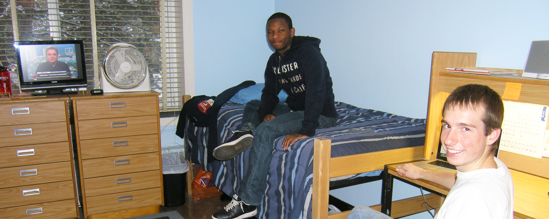 One student sitting in a chair, and another student sitting on the bed in their shared double-standard room, facing the camera