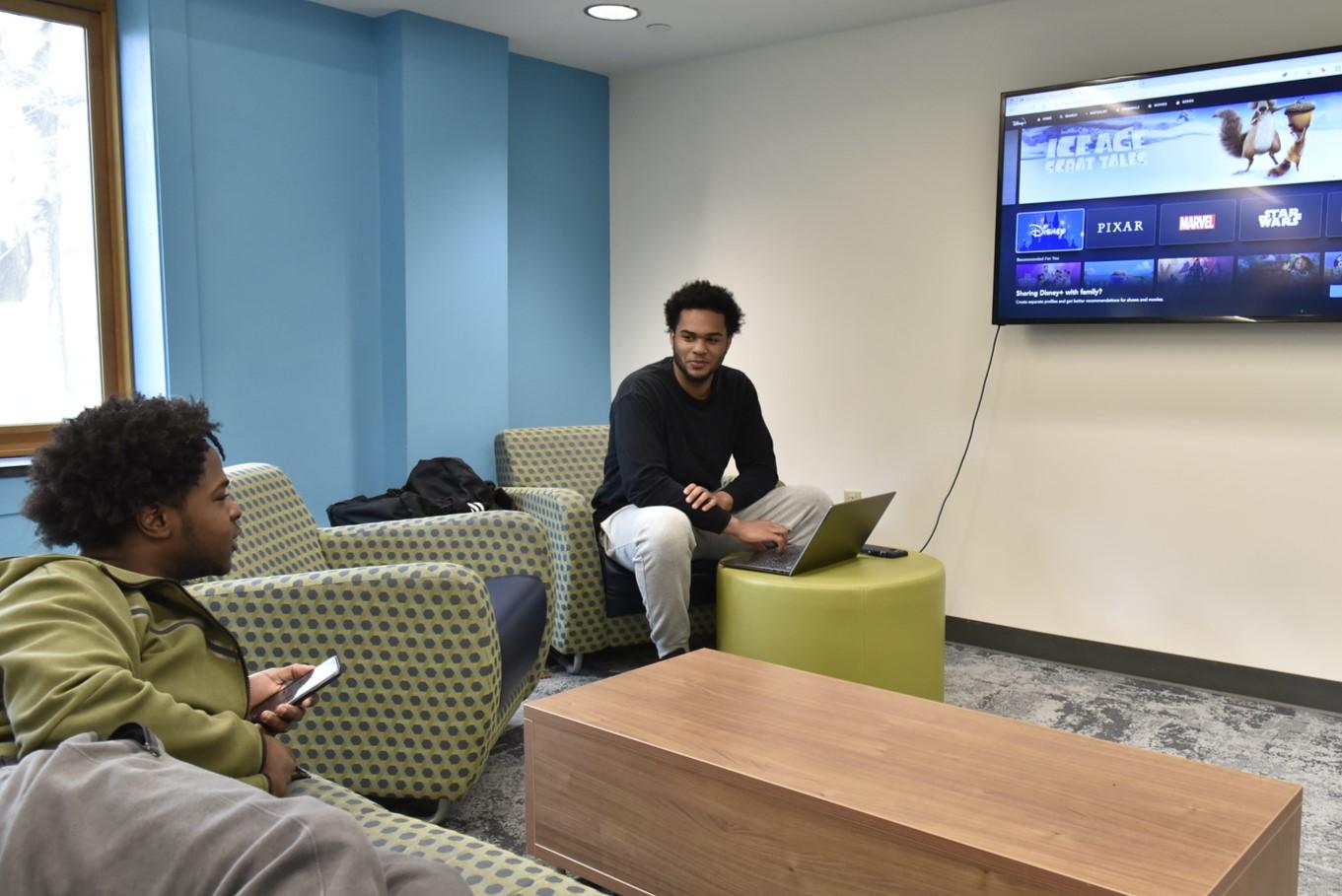A student has their laptop connected to the TV in Scales Hall's basement lounge, talking to another student and deciding what show to stream.