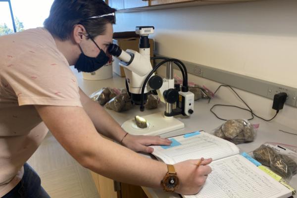 Charles in a laboratory, using a microscope to study a rock and writing notes in a notebook. Charles is wearing a mask.
