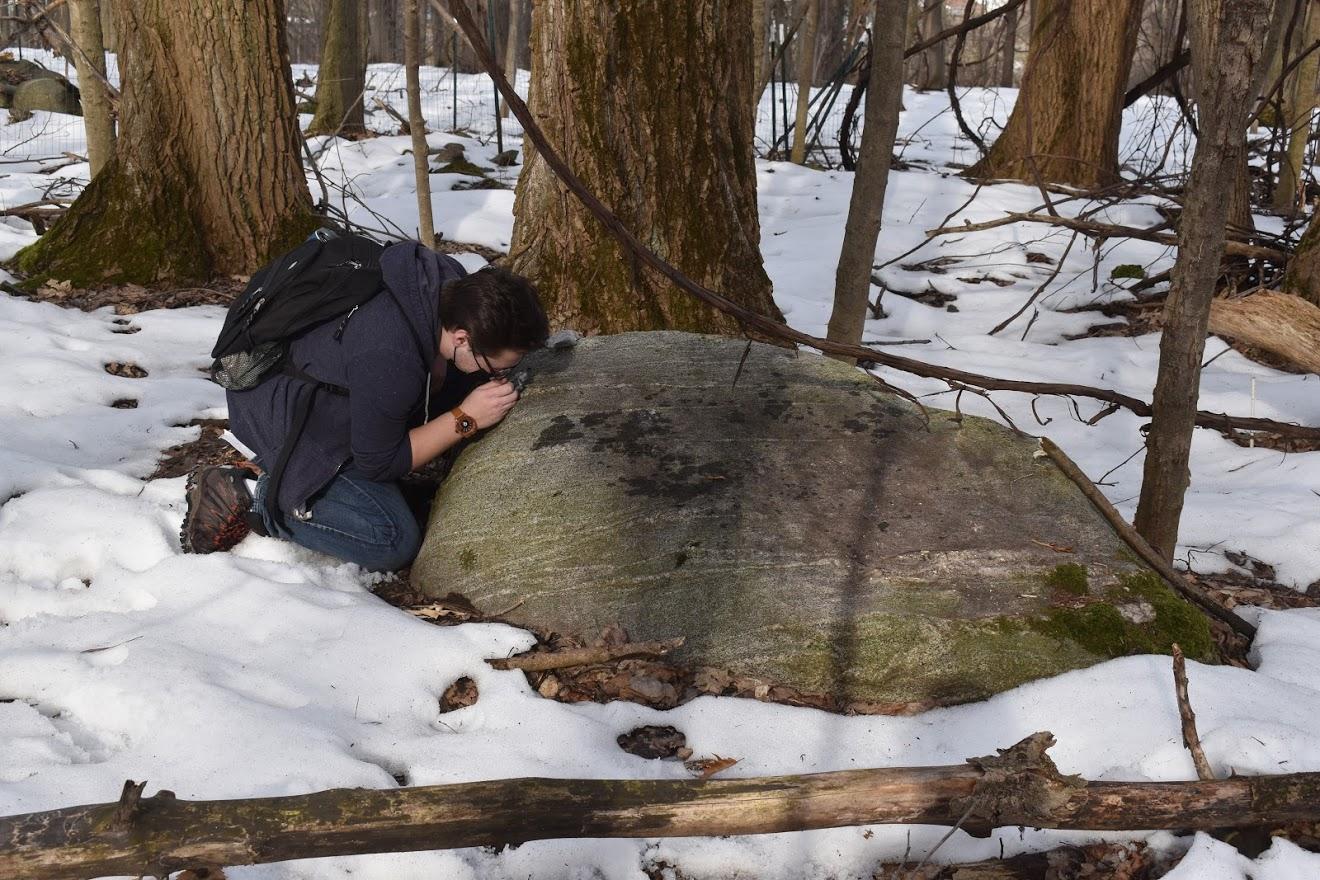 Student kneeling by large boulder in snow, examining the boulder with magnifying lens