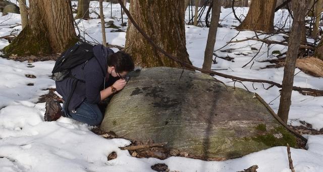 Student kneeling by large boulder in snow, examining the boulder with magnifying lens