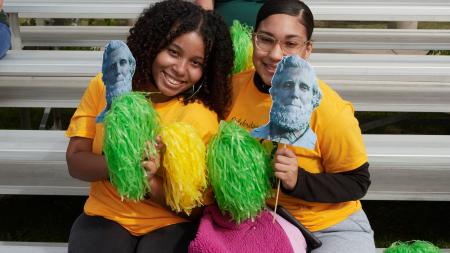 Two students smiling for a photo on a very sunny dau at SUNY Oswego's Green and Gold day. The two students are holding paper cutouts of the Sheldon Statue and holding green and gold pom poms.